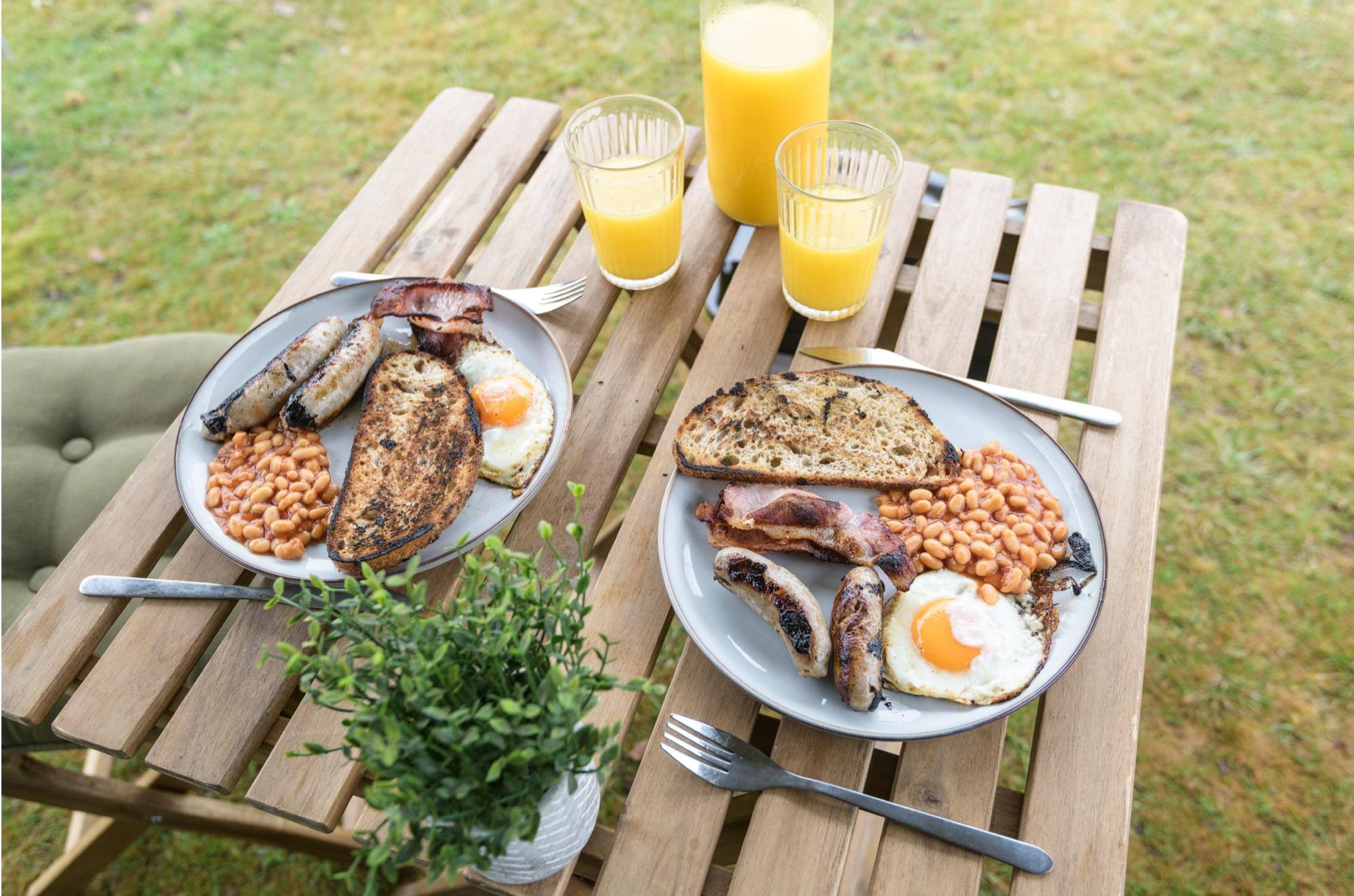 A camping table laden with a two plates of cooked breakfast and two glasses of orange juice