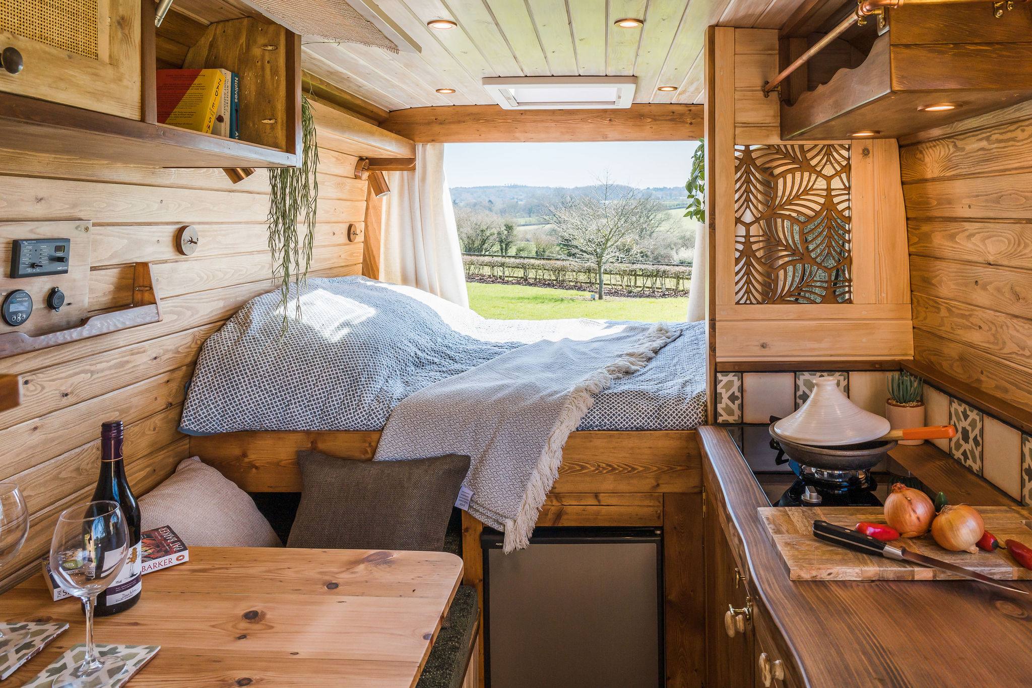 The interior of the campervan showing the rear doors open and he bed. The full interior is cladded in warm tone wood. To the left is the kitchen area