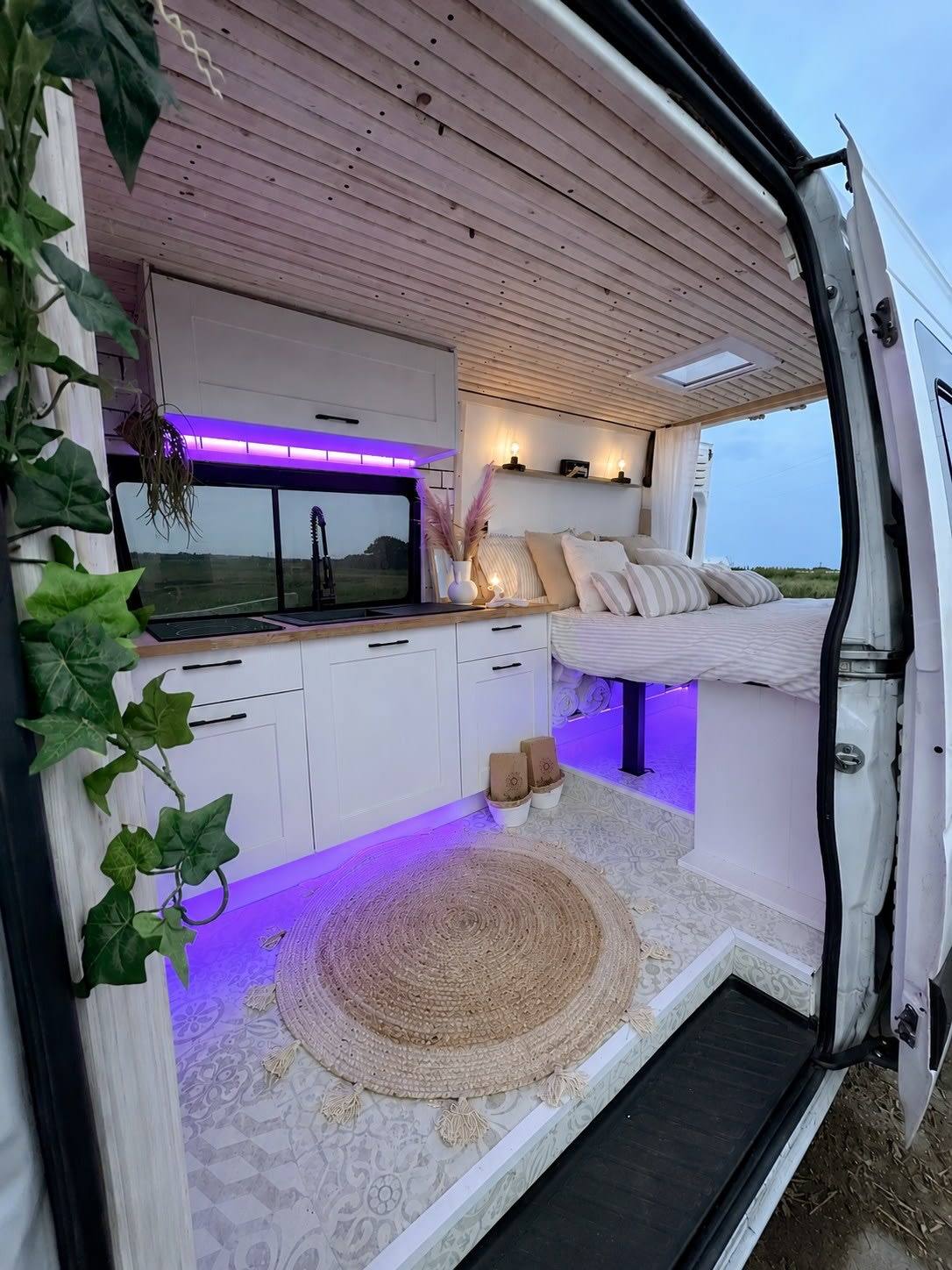 View of a campervan interior through the side door. Inside it has a wood clad cieling, bed and kitchen area all in neutral tones with soft lighting