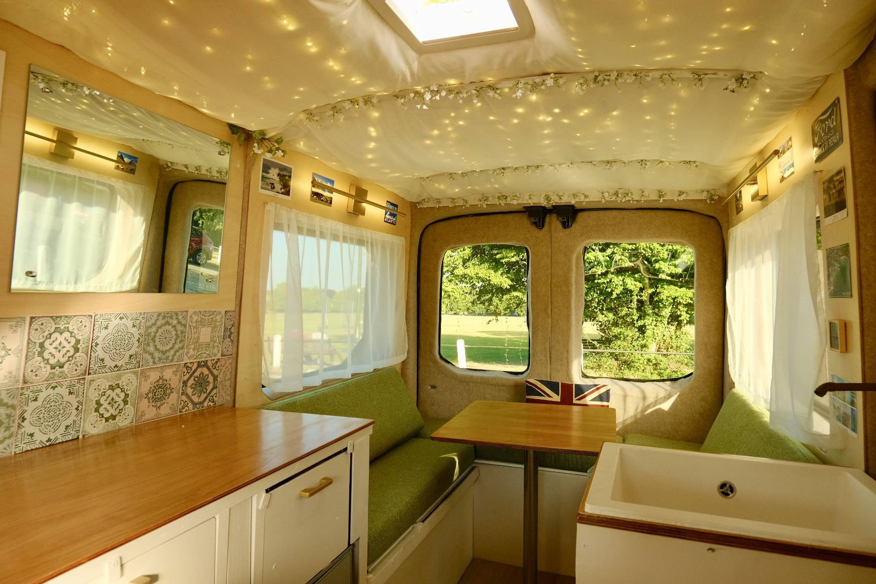 A cozy interior of a camper van with green seating, a wooden table, a small sink, and large windows showing greenery outside. Fairy lights add a starry effect to the ceiling.