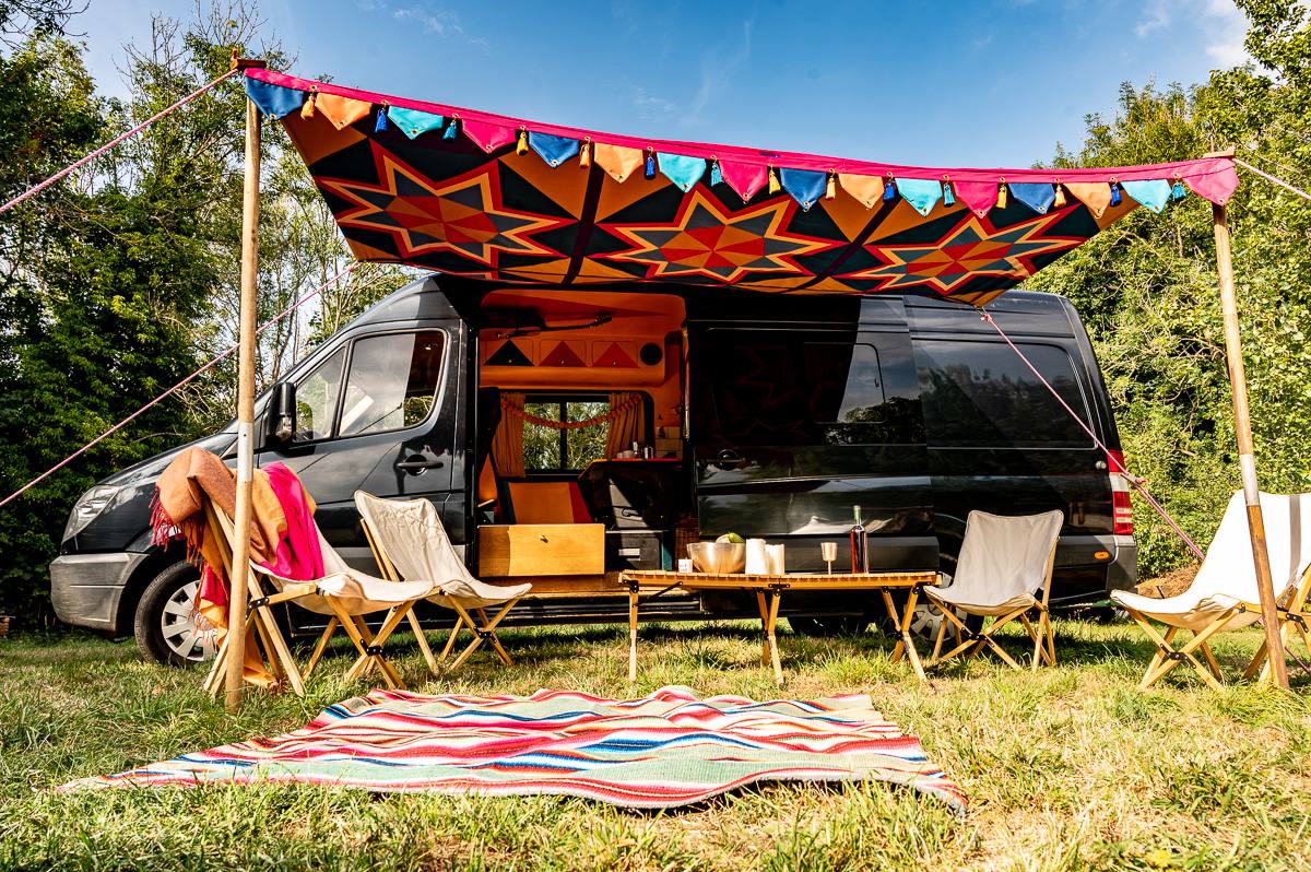 Colorful camper van with patterned awning, bunting, folding chairs, and striped rug in a sunny grassy setting; cheerful outdoor camping scene.