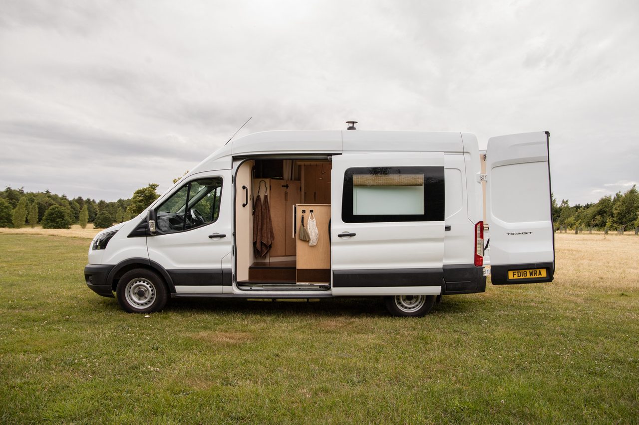 White camper van with open side and rear doors, revealing a cozy wood-panelled interior, parked in a grassy field under cloudy skies.