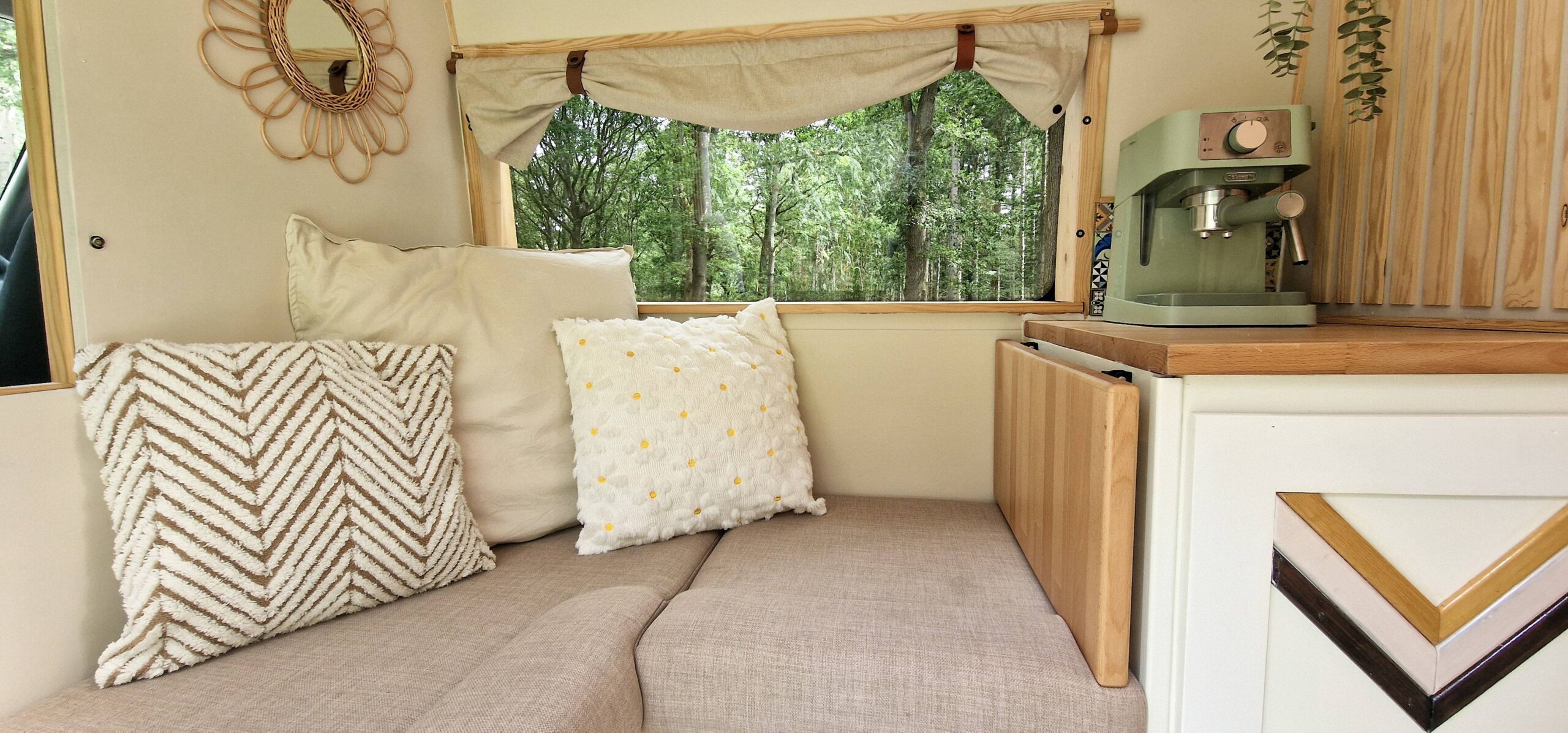 A cozy, well-decorated van interior featuring a small seating area with patterned cushions, wooden accents, and a green coffee machine on a counter. Natural light filters in through a window surrounded by light curtains, and greenery is visible outside.