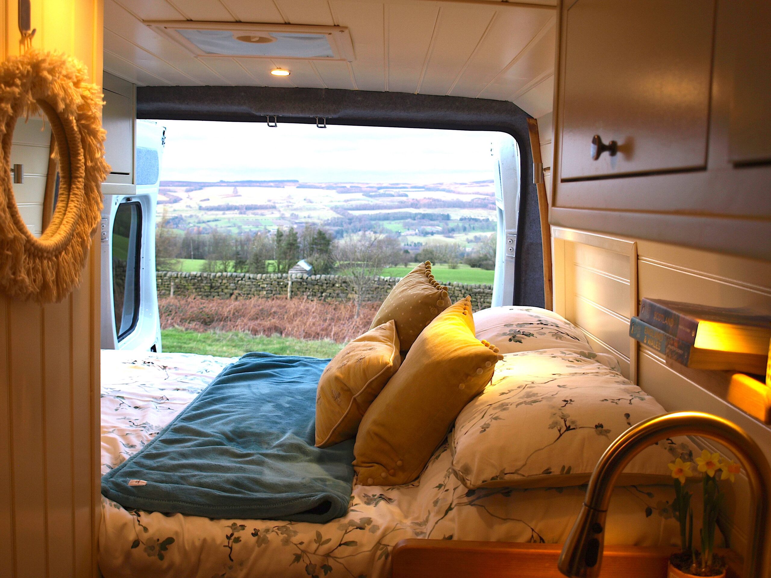 A camper van bedroom with a made bed, pillows, and blanket, opening onto green fields and stone walls.
