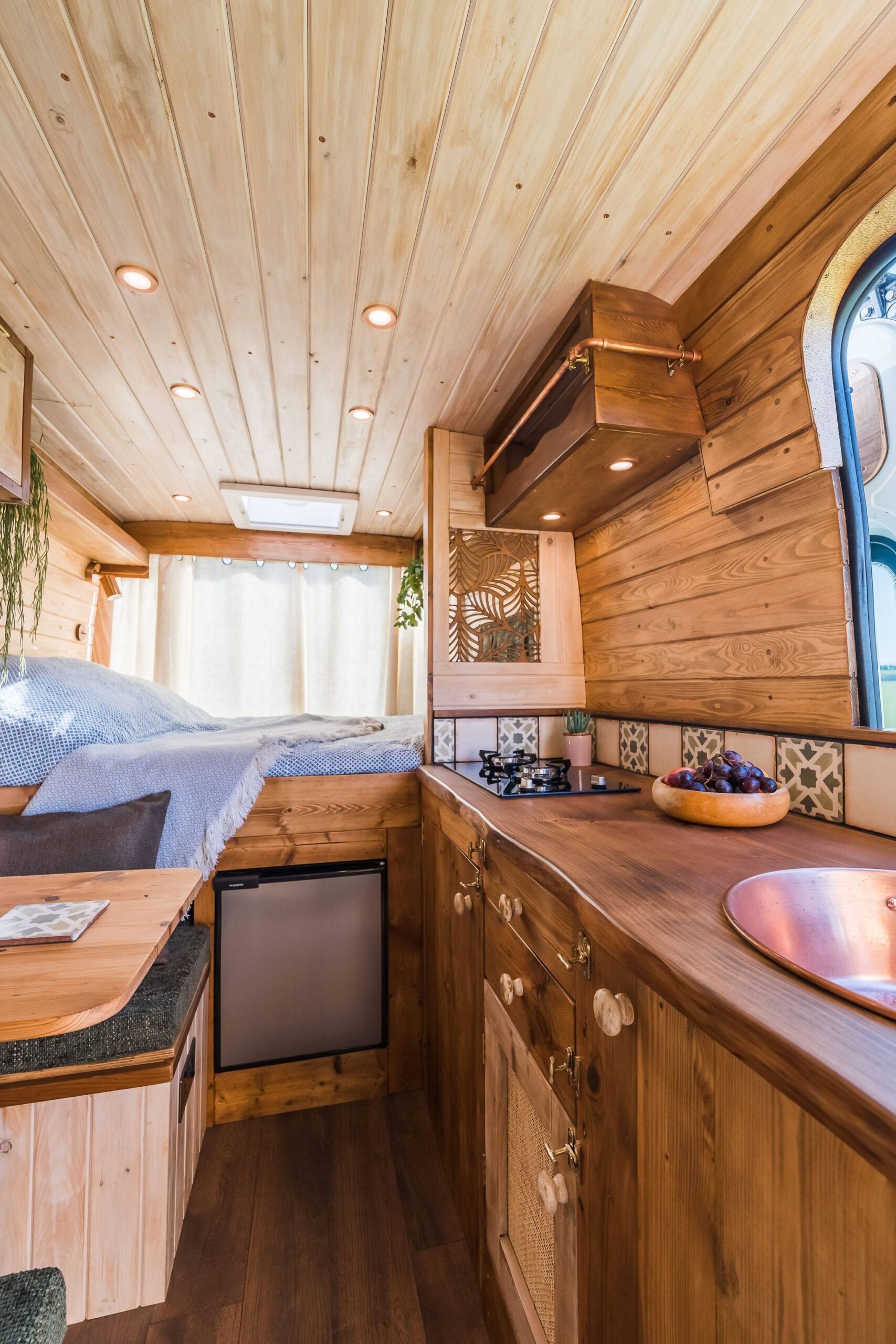 Cozy wood-paneled camper interior with bed, compact kitchen, copper sink, skylight, warm lighting, and rustic bohemian decor.