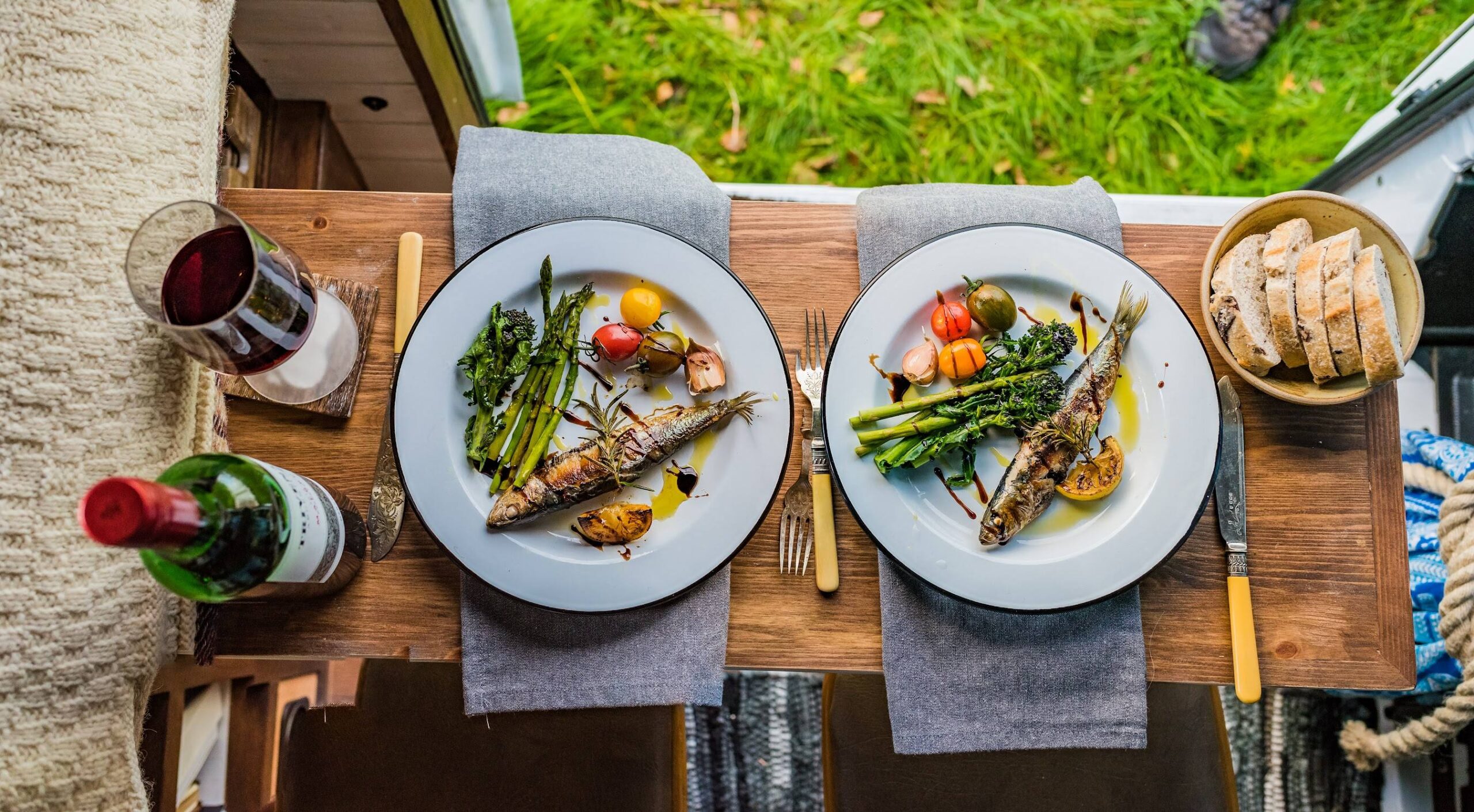 Two plates of grilled fish, asparagus, greens, tomatoes, and lemon sit on a wooden outdoor table with wine, bread, and cutlery.