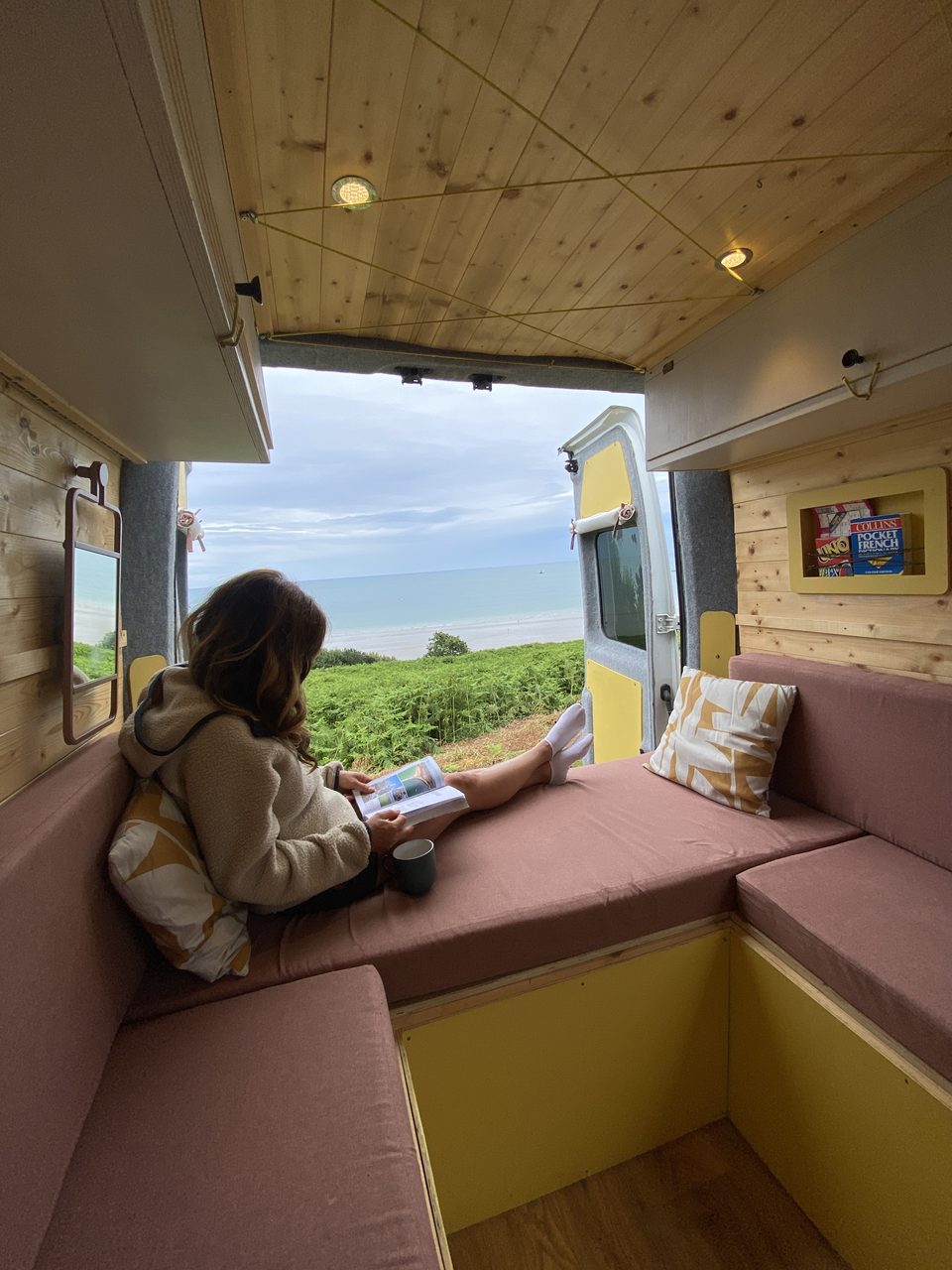 Woman reads in a cozy camper van with pink cushions and a wood ceiling, feet near open doors overlooking lush greenery and the sea.