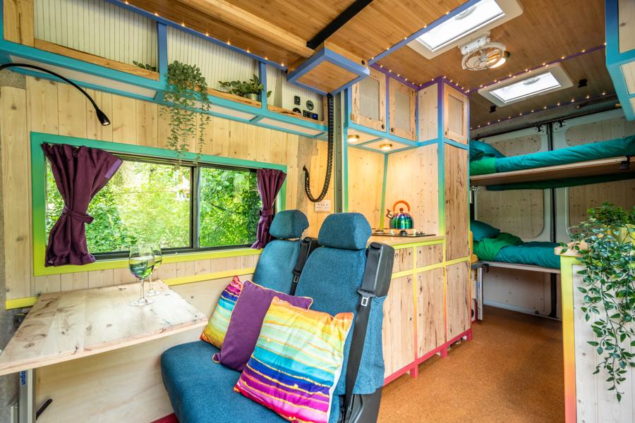 Interior of a colorful campervan featuring a seating area with blue chairs, a table, and decorative pillows, along with a sleeping area in the back and windows providing natural light.
