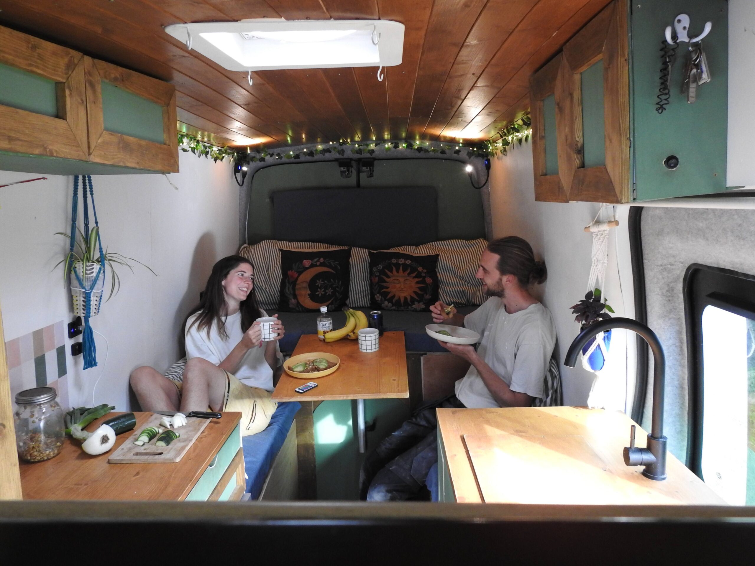 Two people sit facing each other inside a wood-paneled camper van, holding bowls and mugs around a small table with food.
