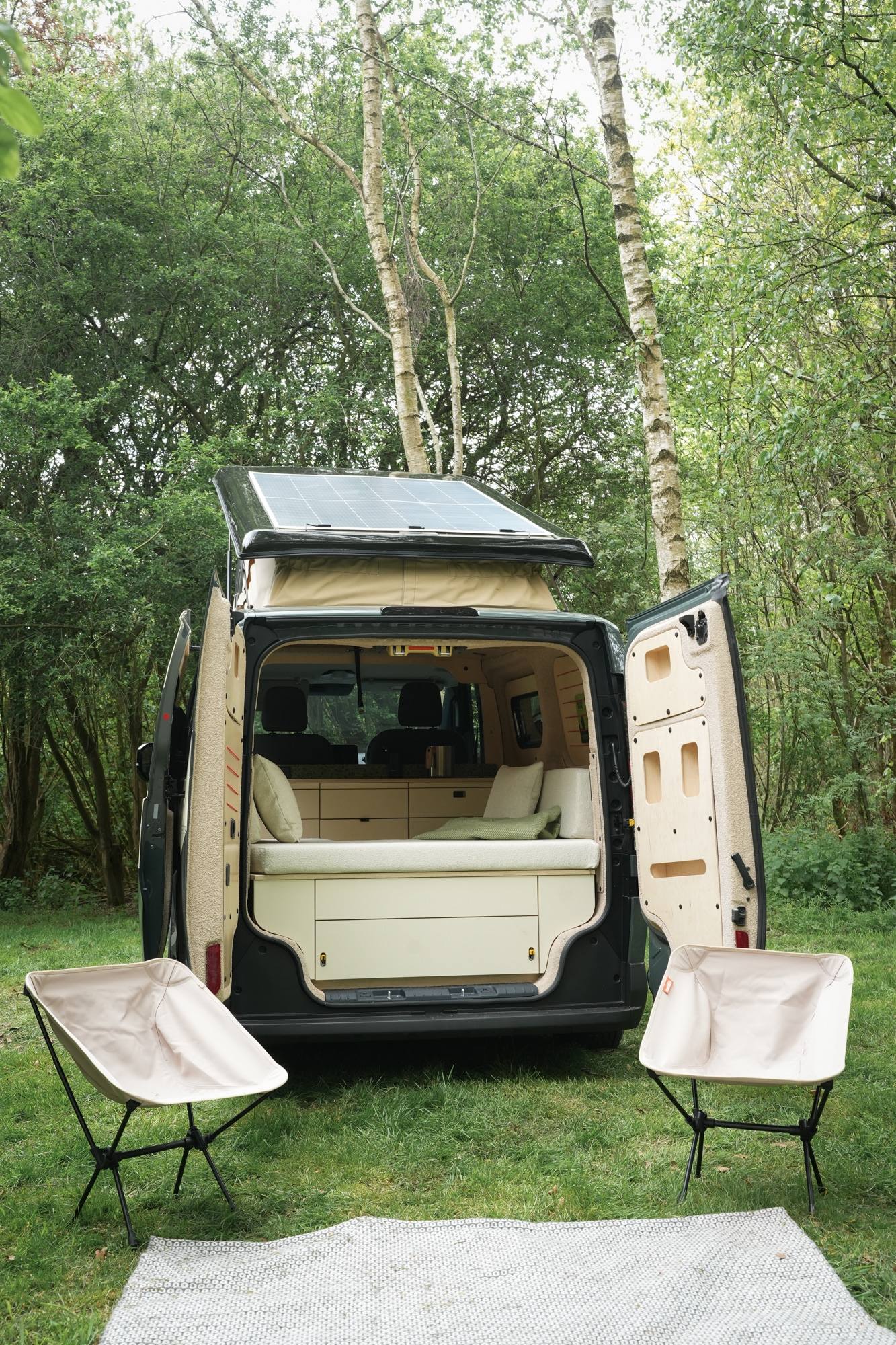 A van parked in the woods, accompanied by a tent and two chairs set up nearby for outdoor relaxation.