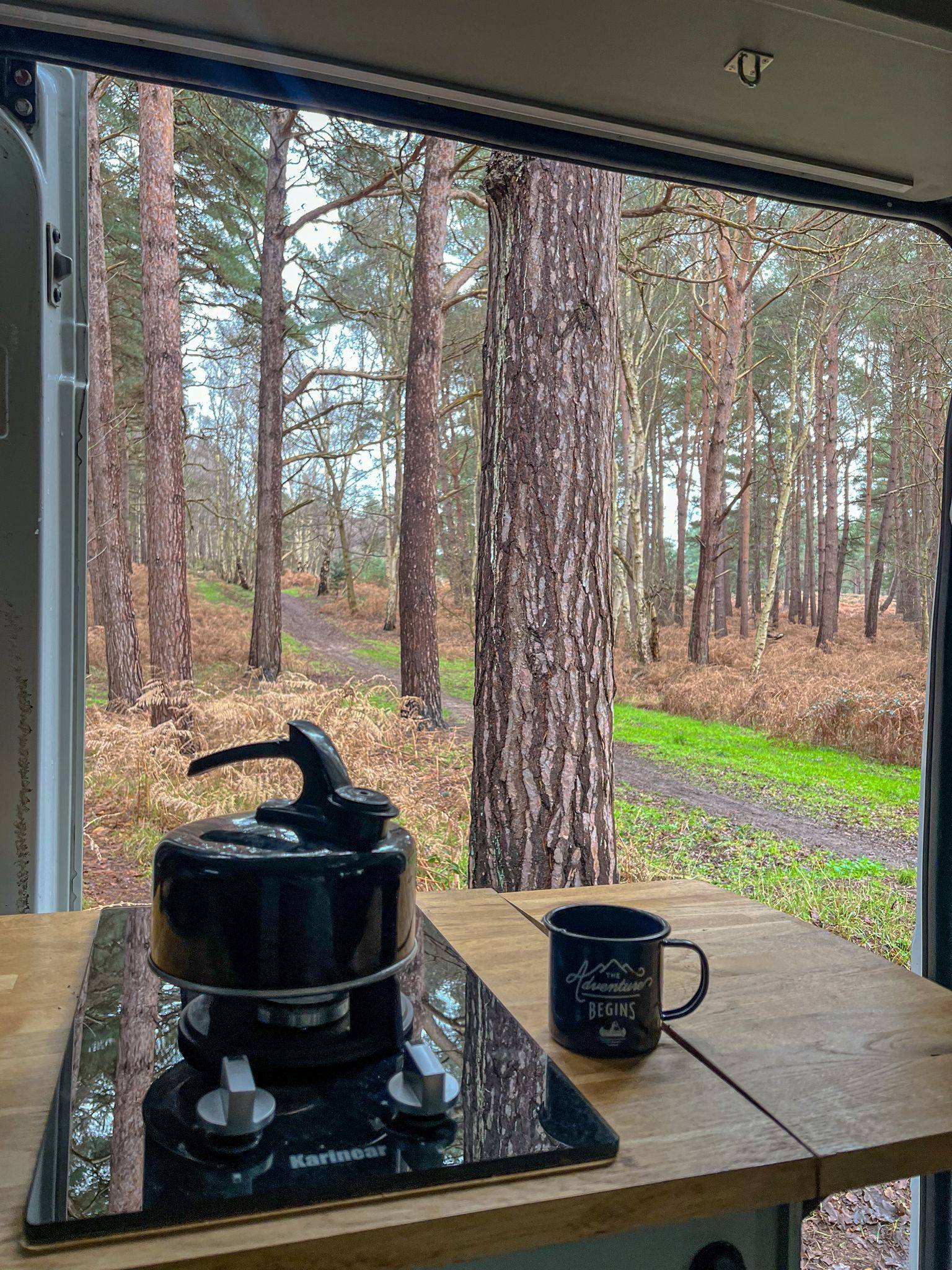 A black kettle and mug reading “Adventure begins” sit on a wooden table overlooking a pine forest through a window.