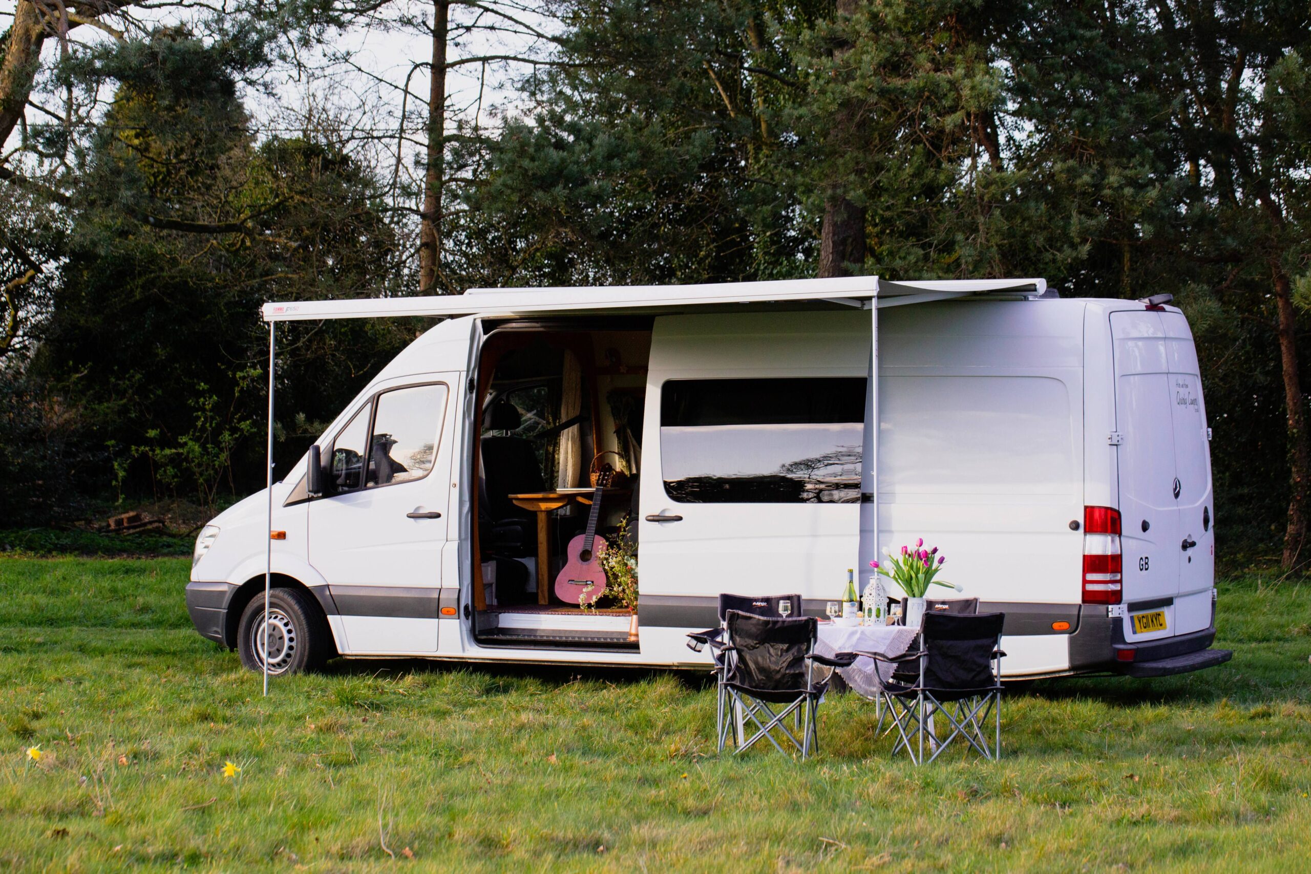 A white camper van parked on green grass, with a guitar visible inside and two folding chairs set up outside on a table with flowers.