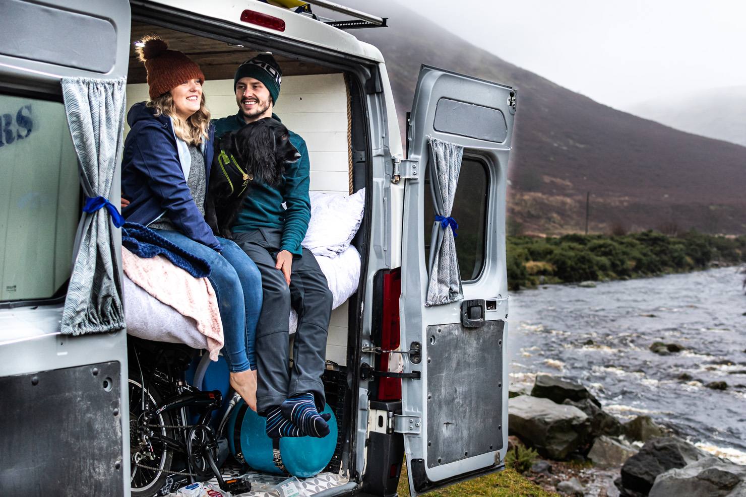 A smiling couple sits with a black dog in the open back of a camper van beside a rocky river, with hills behind them.