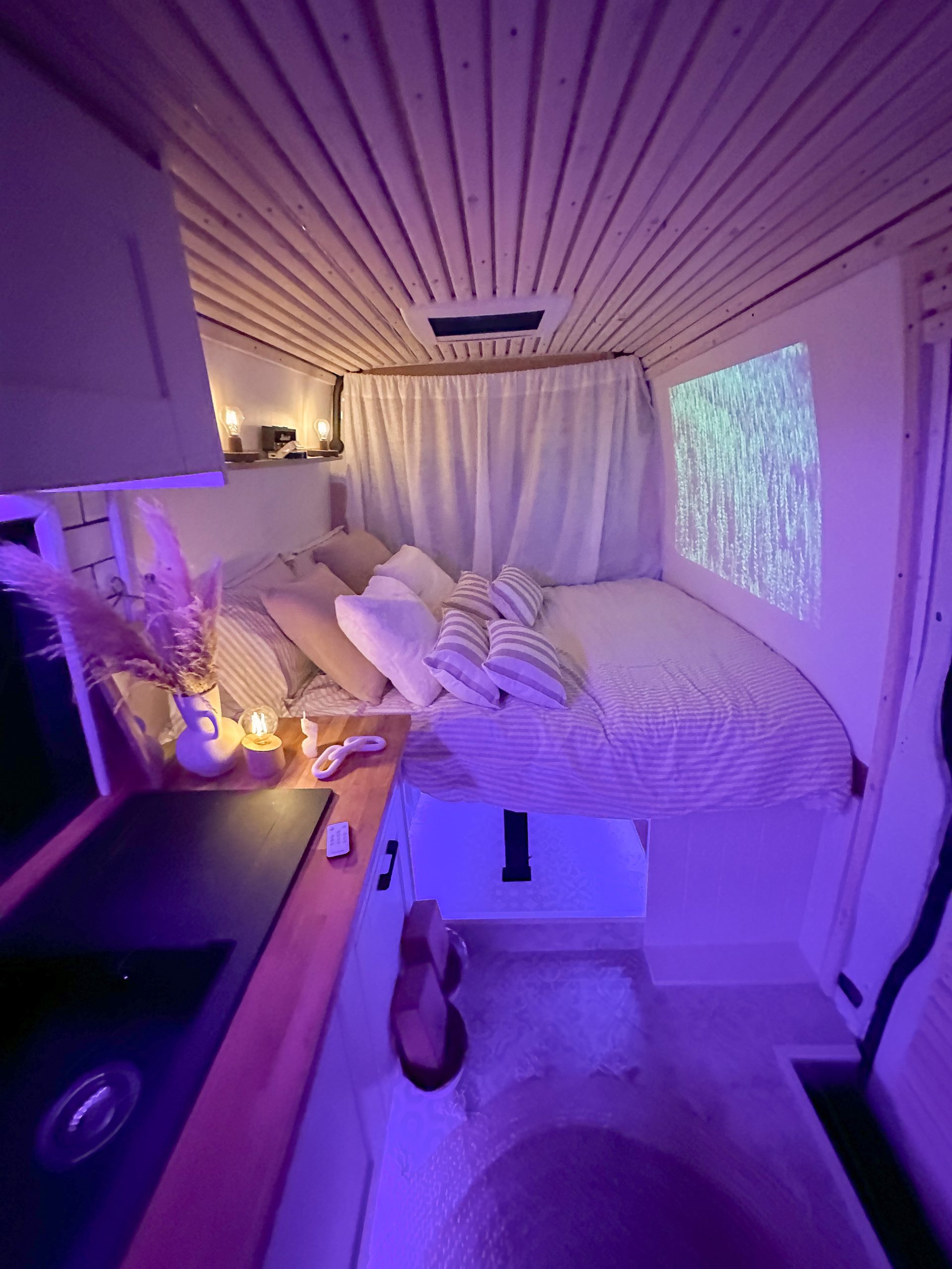 Cozy bedroom area of a campervan. Glowing with purple ambient light, featuring a bed, striped pillows, wood-panelled ceiling, projected wall art, and warm candles.