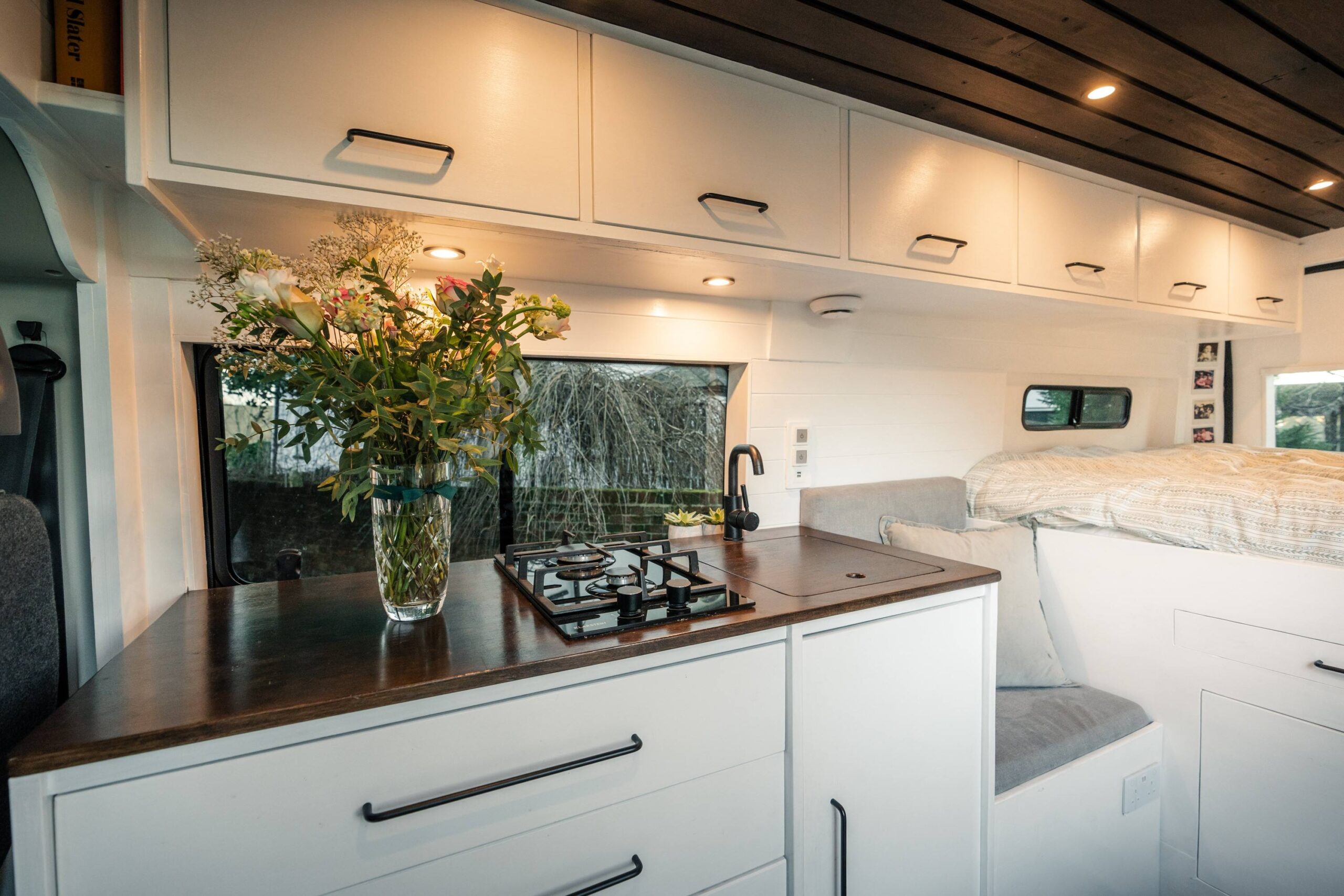 A cozy van interior featuring a kitchen area with a gas stove, a vase of flowers on a countertop, and warm lighting under the cabinets.