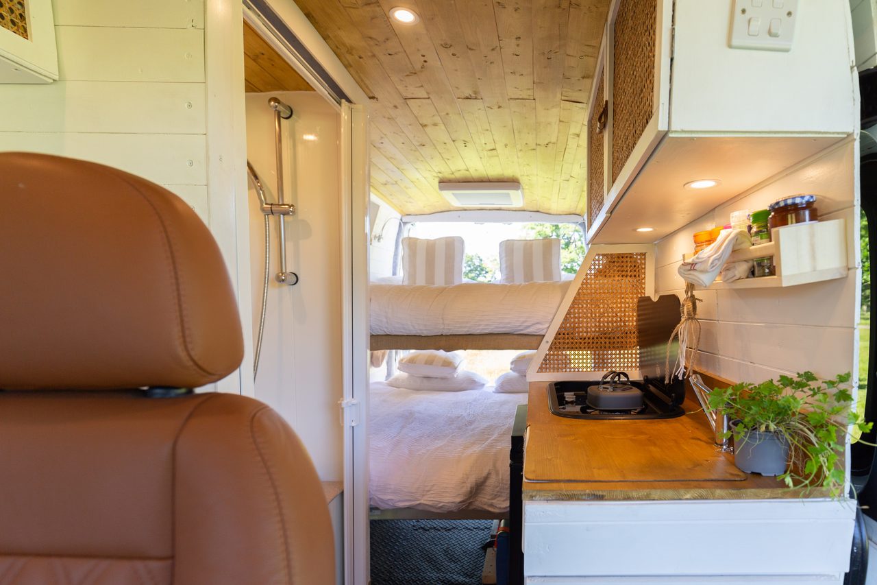 The interior of a campervan has a shower, bunk beds, a kitchenette with shelves, and a potted plant.
