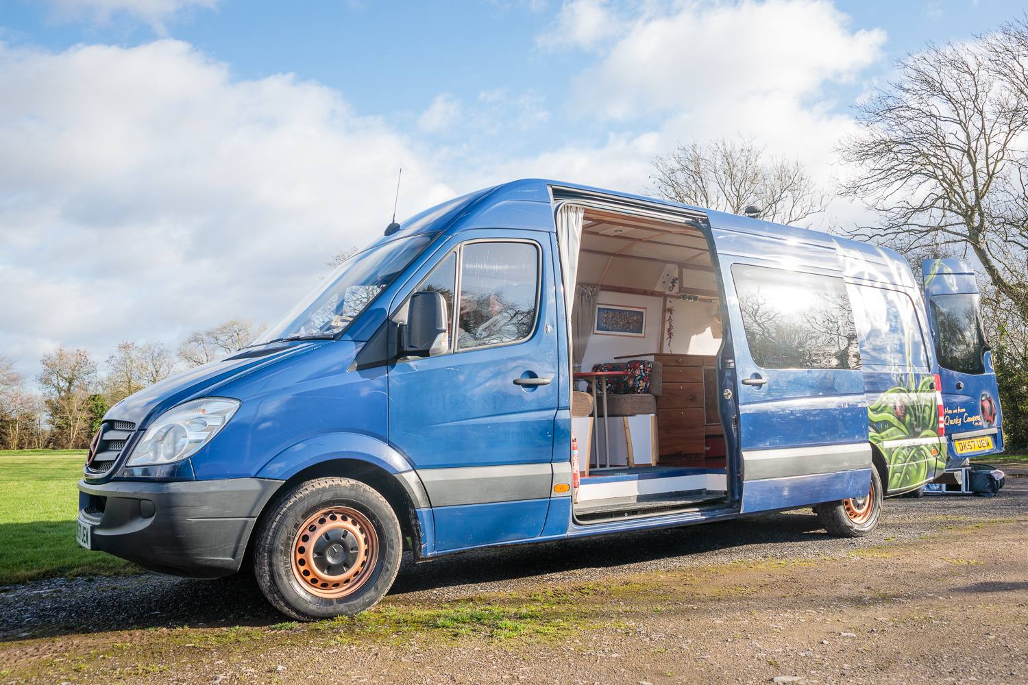 Blue campervan with its side door open, revealing a cozy wood-paneled interior, parked beside a grassy field.