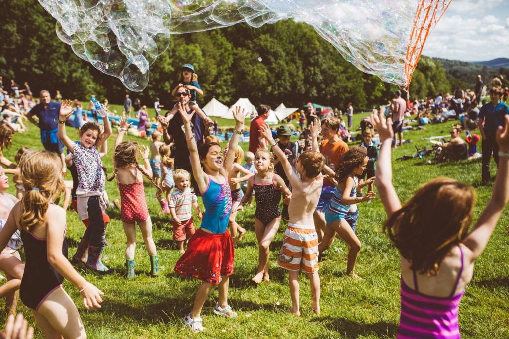 Children reach toward a large sheet of floating soap bubbles at an outdoor gathering, with families and tents on the grassy hillside.