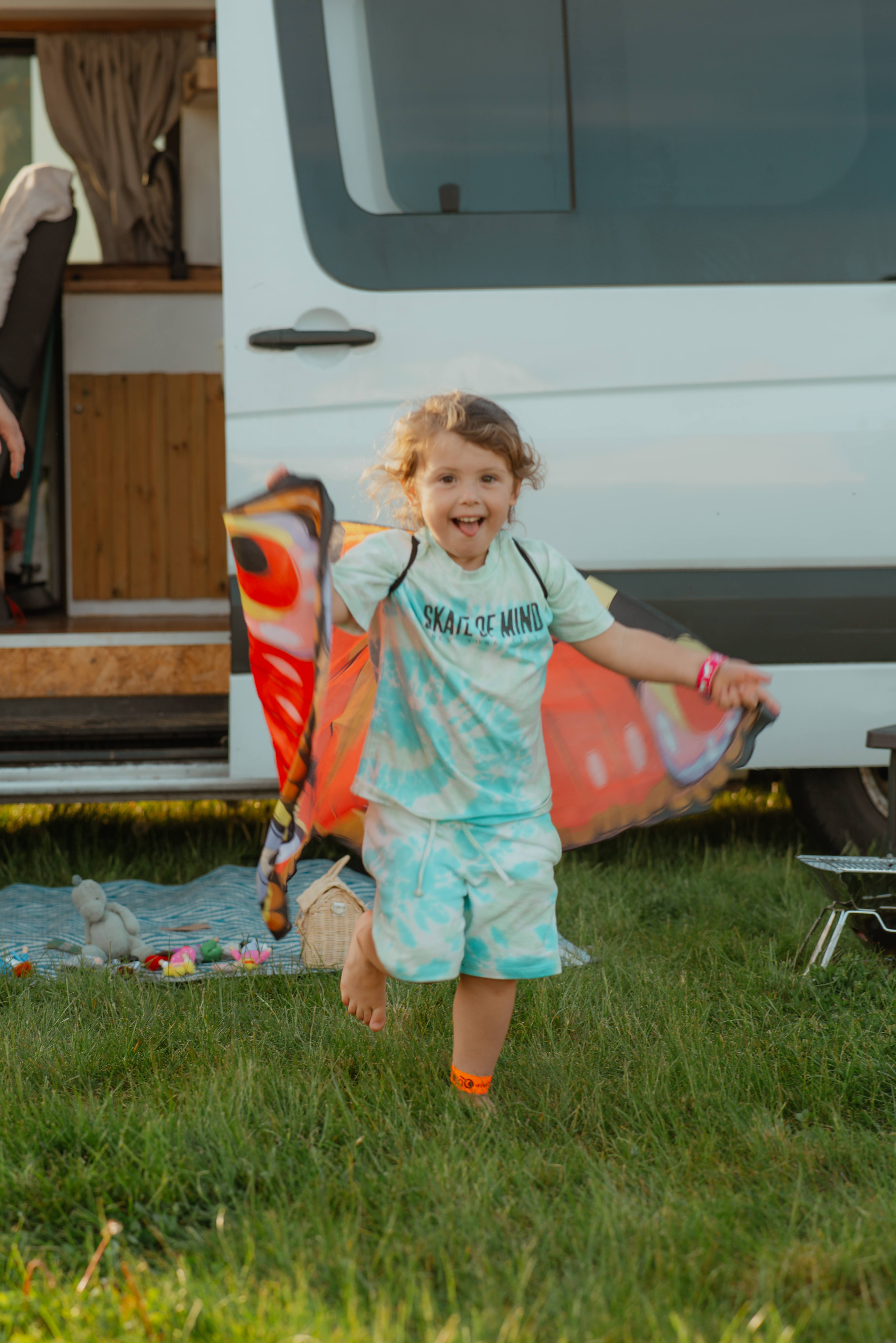 A smiling child runs barefoot on grass, holding out a colorful blanket beside an open campervan.