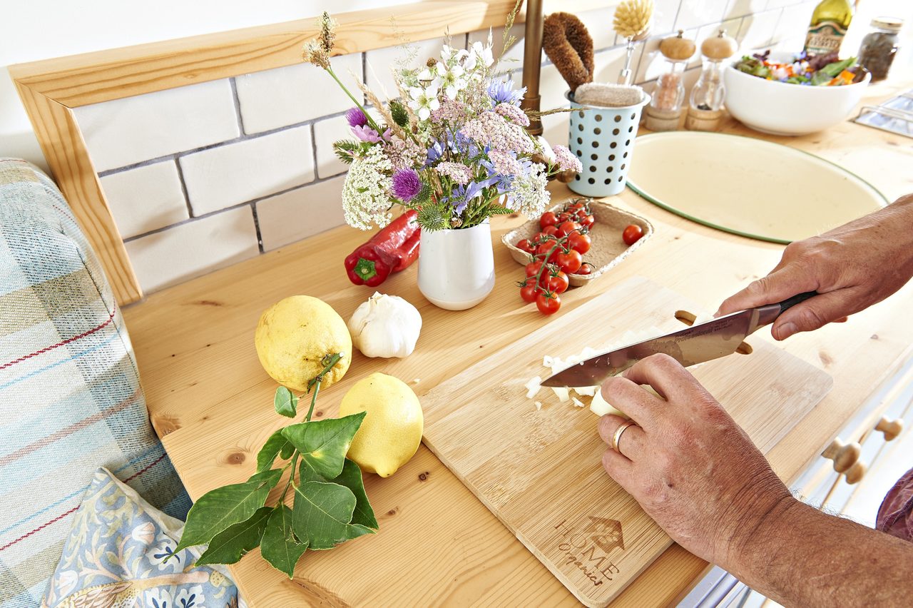 A person cuts fresh food on a wooden cutting board with fruits and vegetables on the kitchen counter.