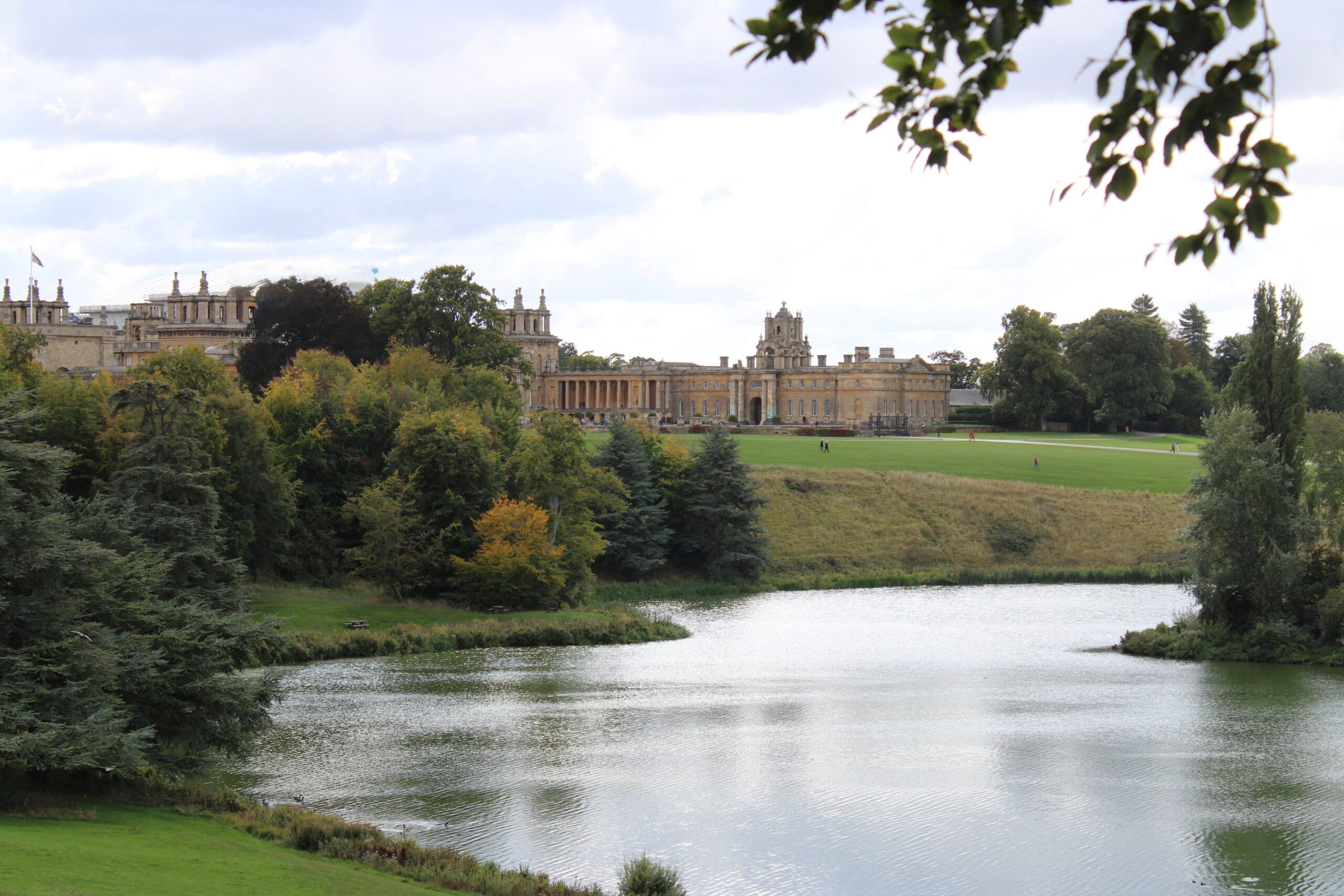Blenheim Palace stands beyond a calm lake and green parkland, framed by trees beneath a cloudy sky, creating a peaceful, grand atmosphere.
