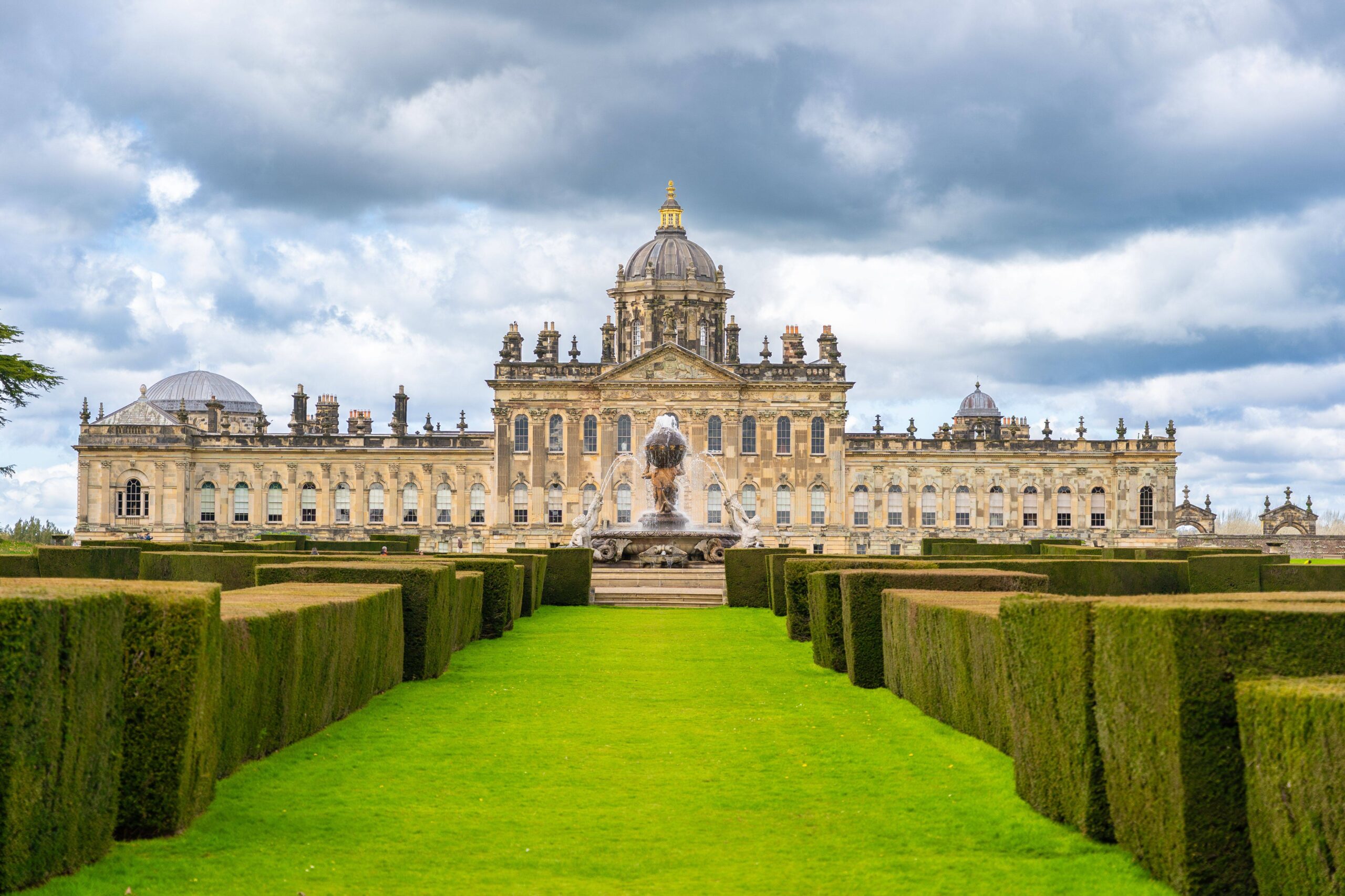 A grand symmetrical palace with ornate stone façades and a central dome stands beyond geometric green hedges beneath a dramatic cloudy sky.
