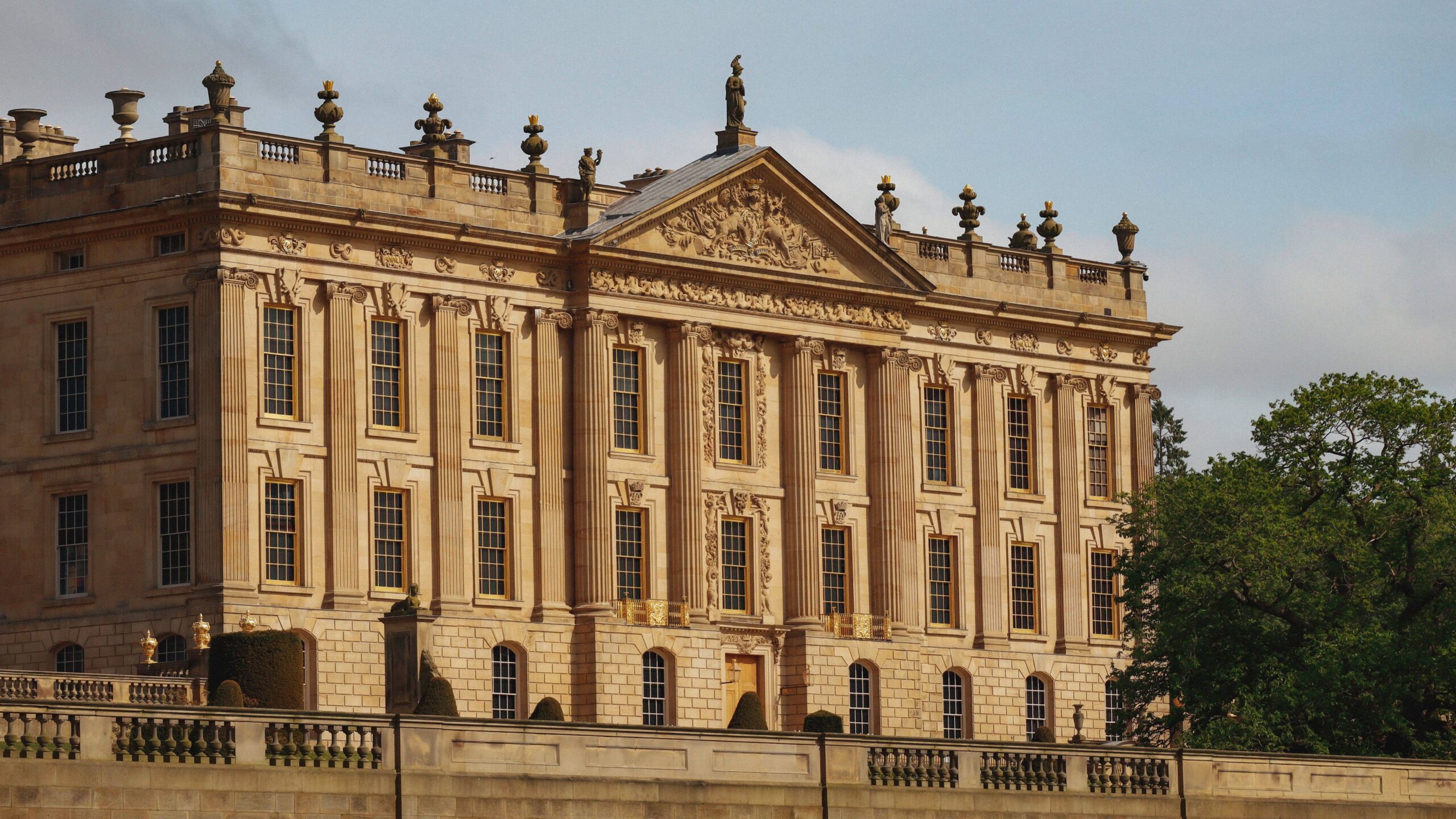 Sunlit neoclassical palace with an ornate stone façade, tall symmetrical windows, rooftop statues, and a balustraded terrace beside green trees.