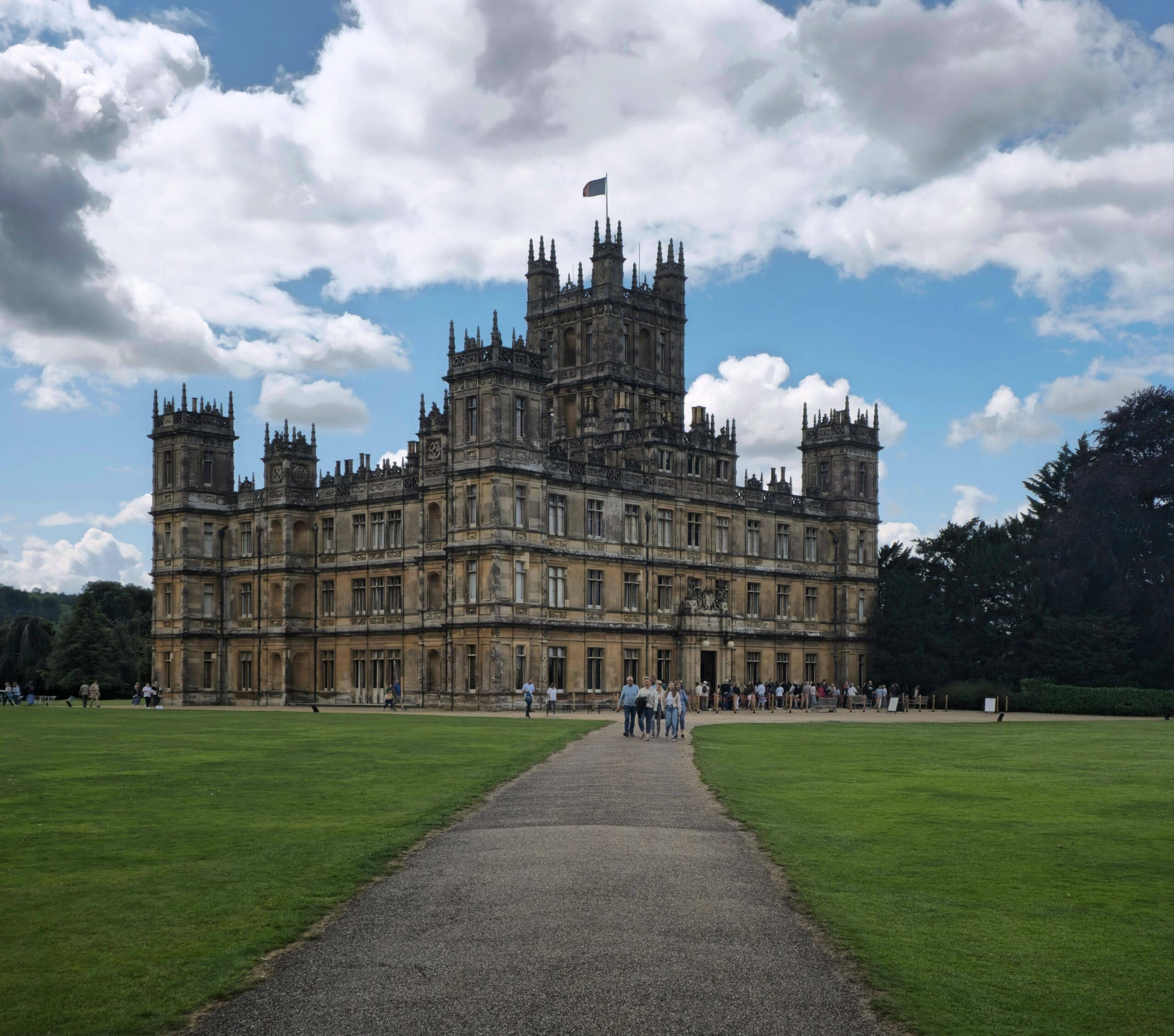 A grand Gothic stone manor rises beyond a gravel path across a green lawn, beneath dramatic clouds; visitors gather at its entrance, creating a stately, historic mood.