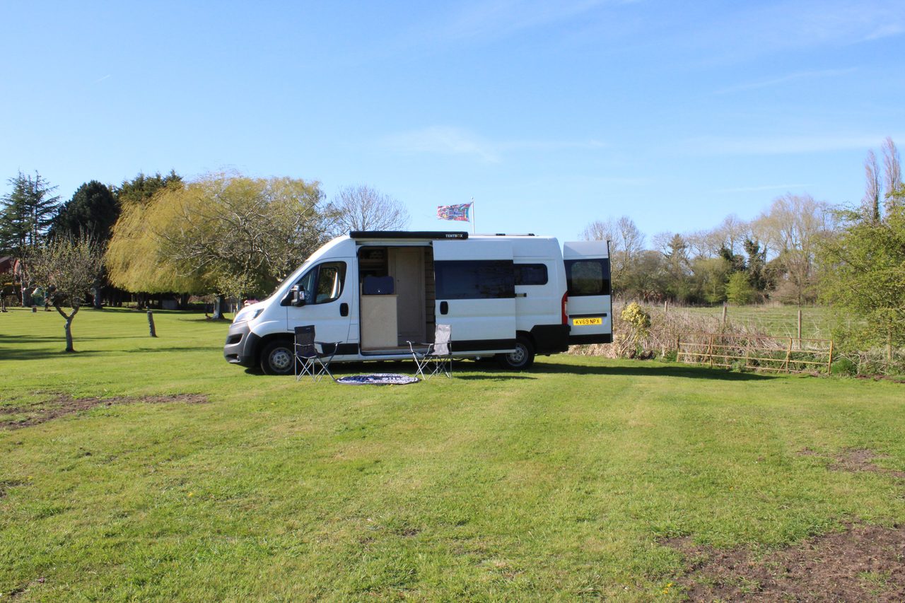 A white campervan with its side door open sits on a sunny green campsite, a small flag flying above it and trees lining the peaceful background.