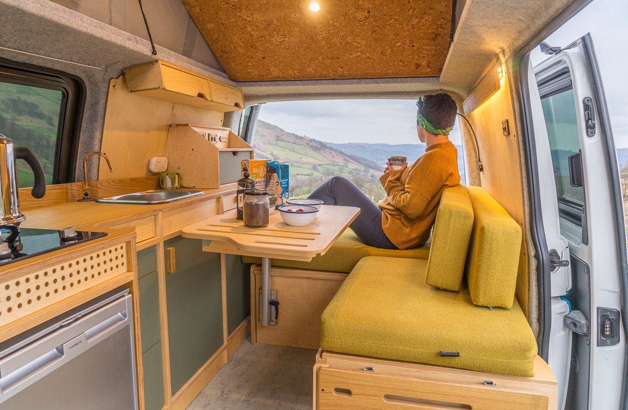 A relaxed traveller in an orange sweater sits in a cozy yellow camper van, gazing at rolling green hills through the open rear, with a compact kitchen nearby.